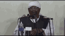 a man wearing glasses and a head scarf stands in front of a microphone