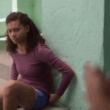 a woman in a purple shirt and blue shorts leans against a wall