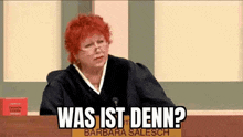 a woman with red hair and glasses is sitting at a table with the words `` was ist denn '' written on it .
