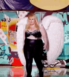 a woman in a black bra and black pants with white wings is standing in front of a graffiti wall .