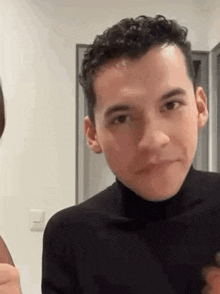 a man with curly hair is wearing a black turtleneck and looking at the camera .
