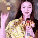 a woman is holding a microphone and waving at the camera while wearing a yellow shirt .