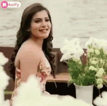 a woman in a floral dress is standing in front of a vase of flowers and smiling .