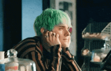 a young man with green hair is sitting at a table with his hands on his face .