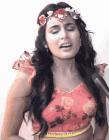 a woman wearing a flower crown and a pink crop top is singing into a microphone