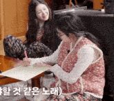 a woman in a pink vest is writing on a piece of paper while another woman looks on