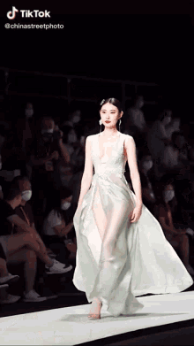 a woman is walking down a runway wearing a white dress with a plunging neckline