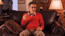 a man in a red sweater sits on a couch holding a bottle of cologne