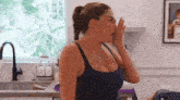 a woman in a blue tank top is covering her mouth with her hand in a kitchen