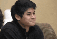 a young man in a black jacket is sitting on a couch and smiling