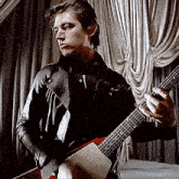 a man in a leather jacket is playing a guitar in front of a curtain