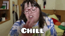a woman wearing glasses and a tie dye shirt is sitting in a bedroom and saying `` chill '' .