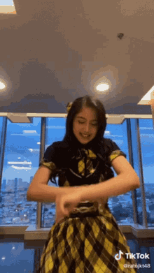 a girl in a yellow and black plaid dress is dancing in front of a window with tiktok written at the bottom