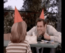 a man and a girl are sitting at a table with a birthday cake . the man is wearing a party hat .