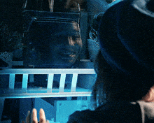 a woman in a blue hat looks at a reflection of a man 's face in a glass