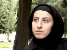 a woman wearing a hijab is standing in front of a tree and looking at the camera .