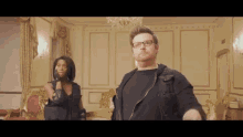 a man and a woman are standing in a room . the man is wearing glasses and a black jacket .