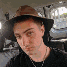 a man wearing a hat and a necklace is sitting in the back seat of a car with a vidu logo in the corner