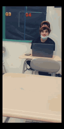 a girl wearing a face mask sits at a desk with a laptop in front of her