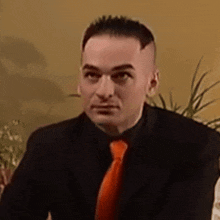 a man wearing a black suit and an orange tie looks at the camera