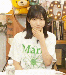 a girl wearing a white t-shirt with a green flower on it is sitting at a table with her hand to her chin .