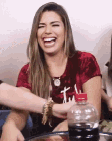 a woman in a red shirt is laughing while sitting at a table with a bottle of soda .