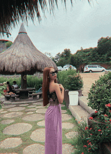 a woman wearing sunglasses and a purple skirt stands in front of a thatched hut