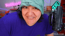 a woman wearing a blue shirt and a green hat holds a camera in front of a neon sign that says nerd craft