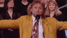 a man in a yellow jacket and tie is standing in front of a microphone in front of a crowd of people .