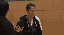 a man in a black hoodie is smiling and waving while another man looks on