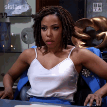 a woman in a white tank top sits at a desk in front of a sign that says " sistas "