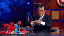 a man in a suit and tie is sitting at a desk in front of a captain america emblem
