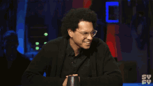 a man wearing glasses and a black shirt is smiling in front of a screen that says syfy