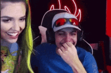 a man and a woman are laughing in front of a computer screen . the man is wearing a hat and sunglasses .