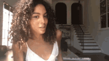 a woman with curly hair in a white tank top stands in front of a staircase