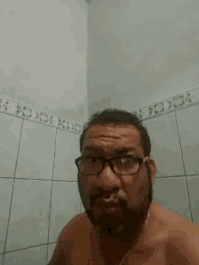 a shirtless man with glasses and a chain around his neck is standing in front of a tiled wall