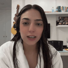 a woman wearing a white robe stands in front of a guitar and a bookshelf