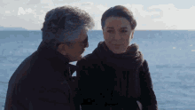a man and a woman are looking at each other in front of the ocean with a hd logo in the corner