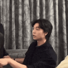 a man in a black shirt is sitting in front of a curtain and talking to someone .