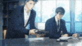two men in suits are sitting at a table and one of them is holding a book .