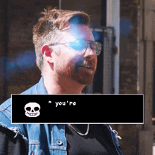 a man wearing glasses and a denim jacket is standing next to a screen that says you 're