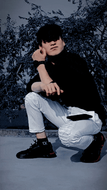a man wearing a black turtleneck and white pants is squatting down with his hand on his chin