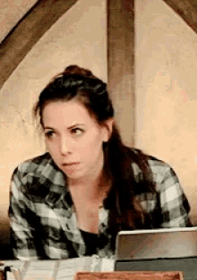 a woman wearing a plaid shirt sits at a table with a laptop
