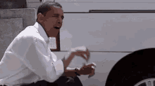 a man in a white shirt is sitting next to a white van and making a funny face .