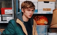 a young man in a green jacket is standing in front of a shelf full of boxes and a pumpkin