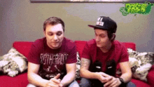 two men are sitting on a red couch and one is wearing a hat that says ny