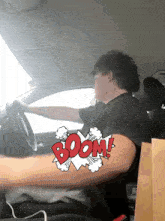 a man in a black shirt is driving a car with a boom graphic on his arm