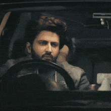a man with a beard is driving a car and covering his mouth with his hand