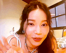 a woman wearing a necklace and earrings is sitting in front of a window and waving .