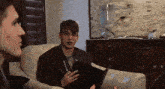 two men are sitting on a couch one is holding a notebook and the other is holding a pen